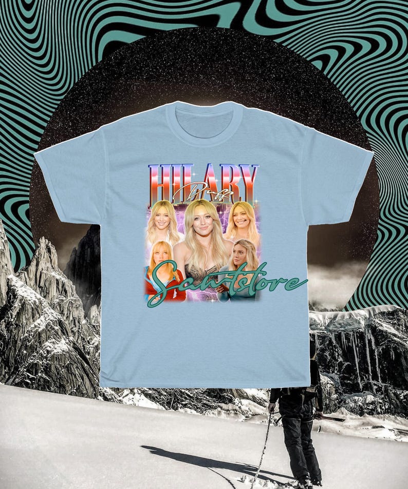 May include: Pale blue t-shirt with a graphic of a woman's image and the name "HILARY" in a retro font. The shirt also features the text "Scents Store" in a cursive font. The background has a retro design.