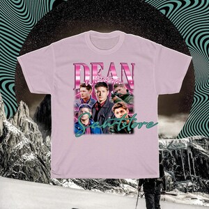 May include: Pink t-shirt with a graphic of Dean Winchester from the TV show Supernatural. The graphic features Dean and other characters from the show in a retro style. The text "Dean Winchester" is in pink and the text "Samstore" is in green.