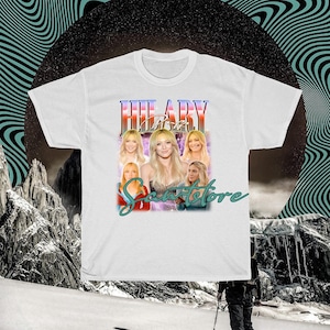 May include: White t-shirt with a graphic design featuring a woman's image and the name "HILARY" in a retro style. The design includes multiple images of the woman and the text "Scents Store". The background has a black circle and wavy lines.