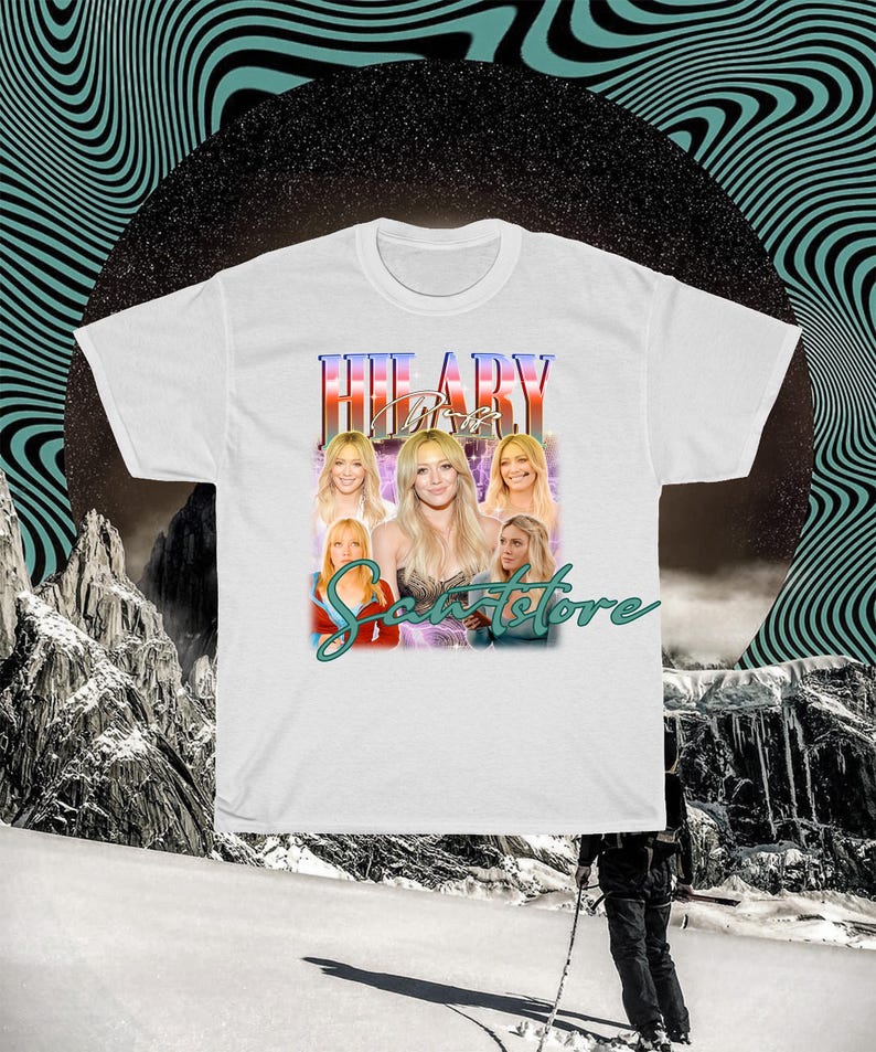 May include: White t-shirt with a graphic design featuring a woman's image and the name "HILARY" in a retro style. The design includes multiple images of the woman and the text "Scents Store". The background has a black circle and wavy lines.