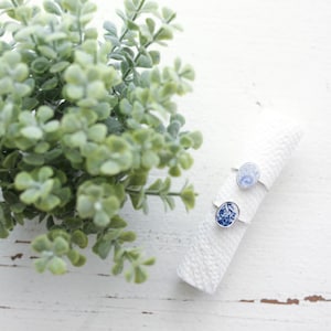 May include: Two oval rings featuring blue and white ceramic-style patterns. One ring displays a light blue marbled design, while the other features a detailed dark blue floral motif. Both rings are set on a white textured cylindrical display.
