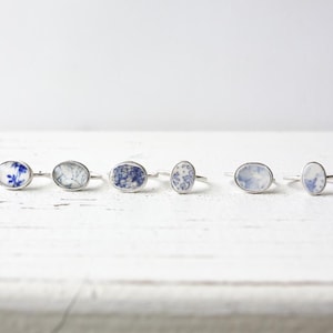 May include: Six silver rings arranged in a row, each featuring an oval-shaped inlay with distinct blue and white floral or patterned designs. The rings have thin, polished metal bands and are displayed in a horizontal line against a plain background.