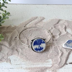 May include: A silver-toned metal bangle bracelet featuring a circular focal point made from a piece of blue and white patterned ceramic pottery. The ceramic insert displays a detailed landscape scene with trees and a figure, set in a metal bezel.