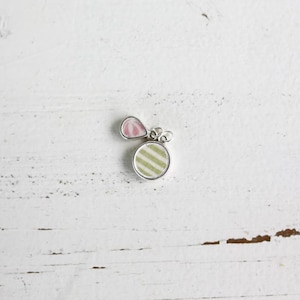 May include: A silver-toned metal charm featuring two distinct components: a small, teardrop-shaped piece with a pink and white pattern and a larger, circular piece displaying a light green and white striped design, both attached to a silver jump ring.