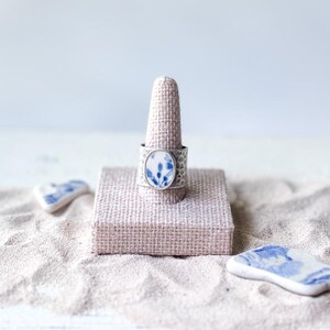 May include: A silver ring featuring a textured band and an oval-shaped ceramic inlay decorated with a blue and white floral pattern. The ring is displayed on a beige fabric cone stand, accompanied by two loose blue and white ceramic shards.