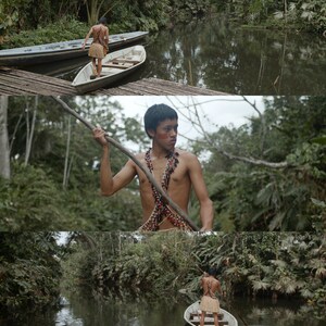 Op de afbeelding: Een jonge persoon in een jungle-omgeving, die een houten kano over een smalle rivier peddelt. De persoon draagt een traditionele kralenketting en een lendendoek.