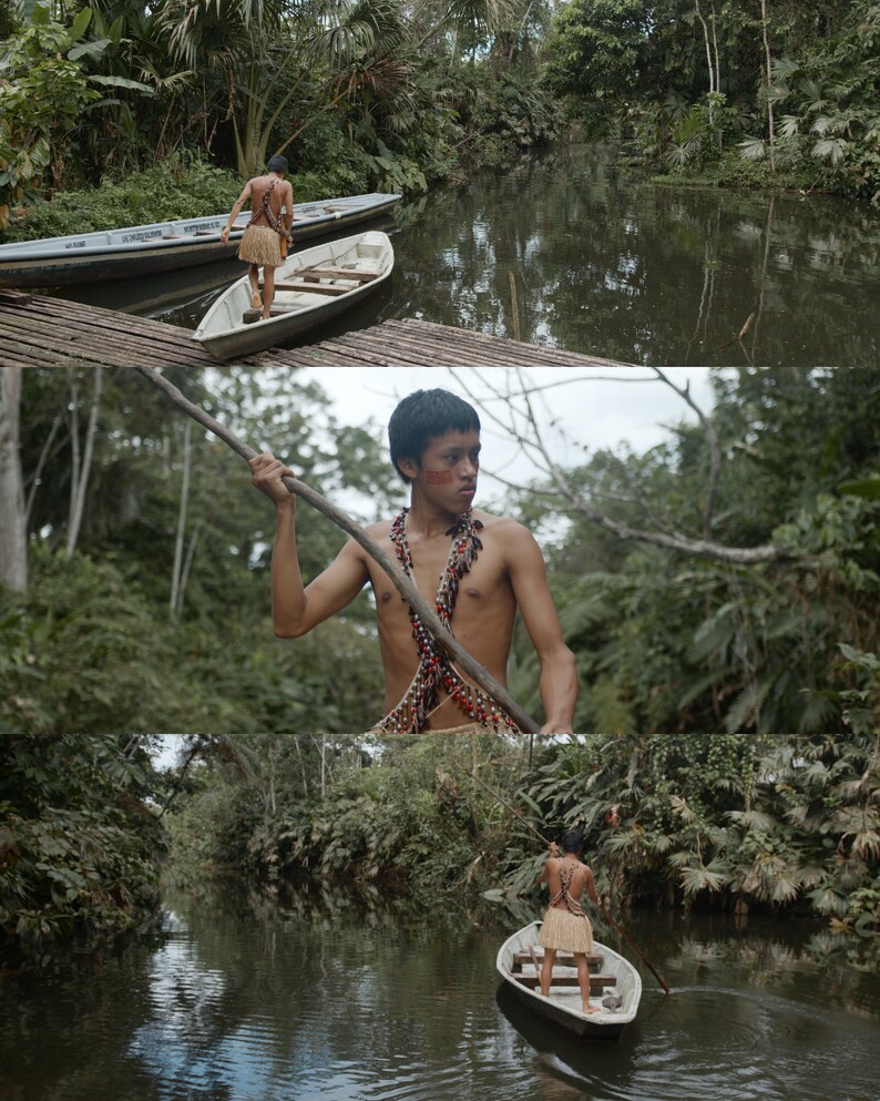 Op de afbeelding: Een jonge persoon in een jungle-omgeving, die een houten kano over een smalle rivier peddelt. De persoon draagt een traditionele kralenketting en een lendendoek.