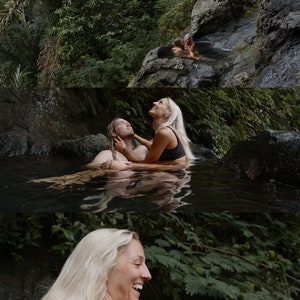 Op de afbeelding: Een stel omarmt elkaar in een natuurlijk zwembad omringd door weelderig groen. De vrouw heeft lang blond haar en draagt een zwarte badpak. De man heeft lang bruin haar en draagt een zwarte badpak. Het stel glimlacht en kijkt elkaar aan.