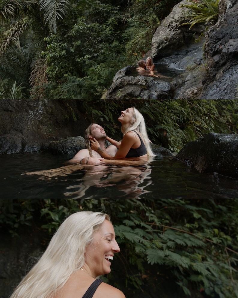 Op de afbeelding: Een stel omarmt elkaar in een natuurlijk zwembad omringd door weelderig groen. De vrouw heeft lang blond haar en draagt een zwarte badpak. De man heeft lang bruin haar en draagt een zwarte badpak. Het stel glimlacht en kijkt elkaar aan.