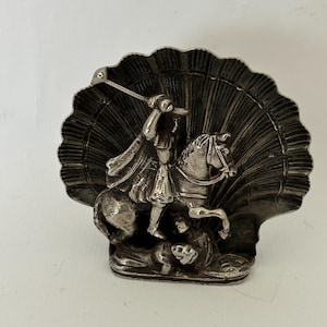 Puede incluir: Figurilla de plata de un caballero a caballo matando a un dragón, sobre un fondo de concha de vieira.