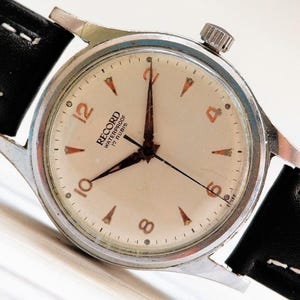 Op de afbeelding: Vintage Record waterdicht horloge met een crèmekleurige wijzerplaat, zwarte wijzers en oranje uurmarkeringen. Het horloge heeft een zilverkleurige kast en een zwarte leren band met witte stiksels. De wijzerplaat toont de tekst "RECORD WATERPROOF 17 RUBIS".
