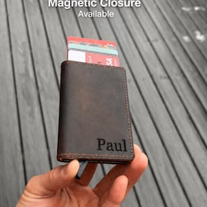 May include: A dark brown leather wallet with visible stitching and the name Paul engraved on the front. The wallet features a card holder mechanism with several cards inserted at the top and includes a magnetic closure feature as indicated by the text.
