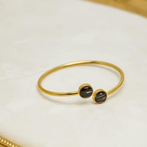 May include: A gold-colored bangle bracelet with two dark, circular accents. The bracelet has a simple, open design. The accents appear to be a dark color with a striped pattern. The bracelet is displayed on a white surface.