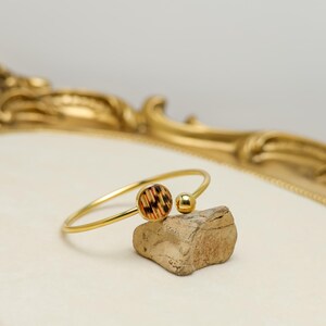 May include: A gold-coloured bangle bracelet with a circular, patterned charm and a small gold ball. The bracelet rests on a small, textured rock. The background features a gold-framed, ornate mirror.