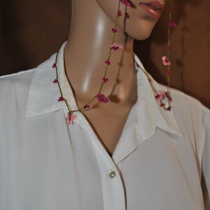 Peut inclure: Un collier et une chaîne de lunettes délicats avec des accents de fleurs roses. Le collier et la chaîne sont enfilés sur un cordon vert. Le modèle porte une chemise blanche boutonnée.