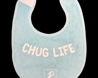 May include: A light blue baby bib with the words "CHUG LIFE" in white block letters. The bib has a light blue trim and a pink velcro closure at the neck. The bottom of the bib has a white square with the letter "P" and the words "PAIGE MARIE".