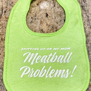 May include: A bright green baby bib with white text that reads "Spitting up on my mom" and "Meatball Problems!". The bib has a rounded shape with a neck opening and is trimmed with a darker green border.
