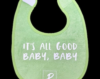 May include: A light green baby bib with the text "IT'S ALL GOOD BABY, BABY" in white. The bib has a rounded shape with a white closure at the neck and a logo at the bottom. The bib is made of a soft, absorbent material.