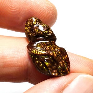May include: A polished, irregularly shaped gemstone with a rich, dark brown base color. The surface is covered in a pattern of golden and iridescent speckles, creating a shimmering effect. The gemstone is held between fingers.