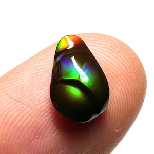 May include: A polished, teardrop-shaped gemstone with a vibrant display of colors. The gem features a dark base with iridescent hues of green, blue, and a flash of red and yellow at the top. The stone is held between two fingers.