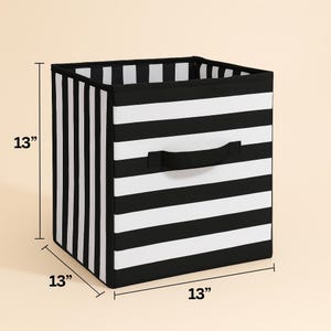 Peut inclure: Cube de rangement à rayures noires et blanches, mesurant 33 cm de chaque côté. Le cube est doté d'une poignée noire sur un côté pour faciliter le transport. Le design présente des rayures horizontales et verticales.