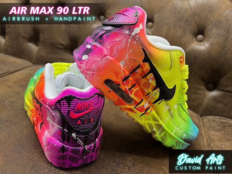 May include: A pair of custom-painted Air Max 90 LTR sneakers with a vibrant rainbow and neon color scheme. The shoes feature a black Nike swoosh and black accents. The shoes are painted with a splatter effect and have a drip effect on the back.