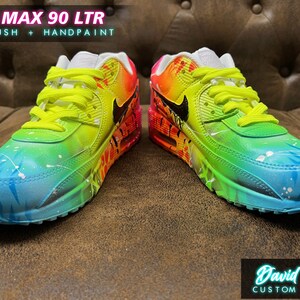 May include: Custom painted Air Max 90 LTR sneakers with a rainbow gradient design. The shoes feature a blue, green, yellow, orange, and pink color scheme with white accents. The Nike swoosh logo is black. The text "AIR MAX 90 LTR" and "AIRBRUSH + HANDPAINT" is visible on the image.