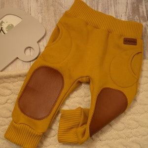 Könnte beinhalten: Senfgelbe Babyhose mit braunen Kniepatches aus Kunstleder. Die Hose hat einen gerippten Bund und Bündchen. Ein kleines braunes Etikett mit Text ist hinten angenäht. Die Hose liegt auf einer cremefarbenen Strickdecke.
