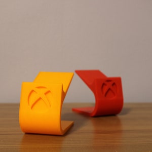 May include: Two 3D printed Xbox controller stands, one orange and one red, with the Xbox logo on the front. The stands are on a wooden surface.