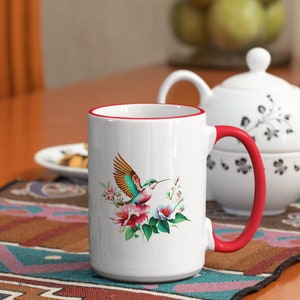 Könnte beinhalten: Weiße Keramiktasse mit rotem Rand und Henkel. Die Tasse zeigt einen bunten Kolibri mit Blumen auf weißem Hintergrund.