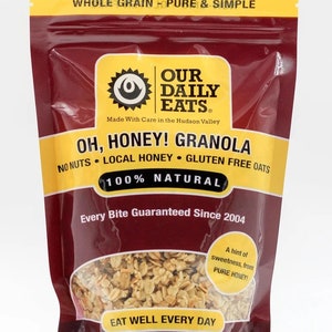 Puede incluir: Una bolsa roja, amarilla y blanca de Our Daily Eats Oh, Honey! Granola. La bolsa presenta un sol con una cara y el texto "Our Daily Eats" en un c&iacute;rculo amarillo. La bolsa tambi&eacute;n presenta el texto "Oh, Honey! Granola" en letras grandes y negritas. La bolsa es de 12 onzas (340 gramos).