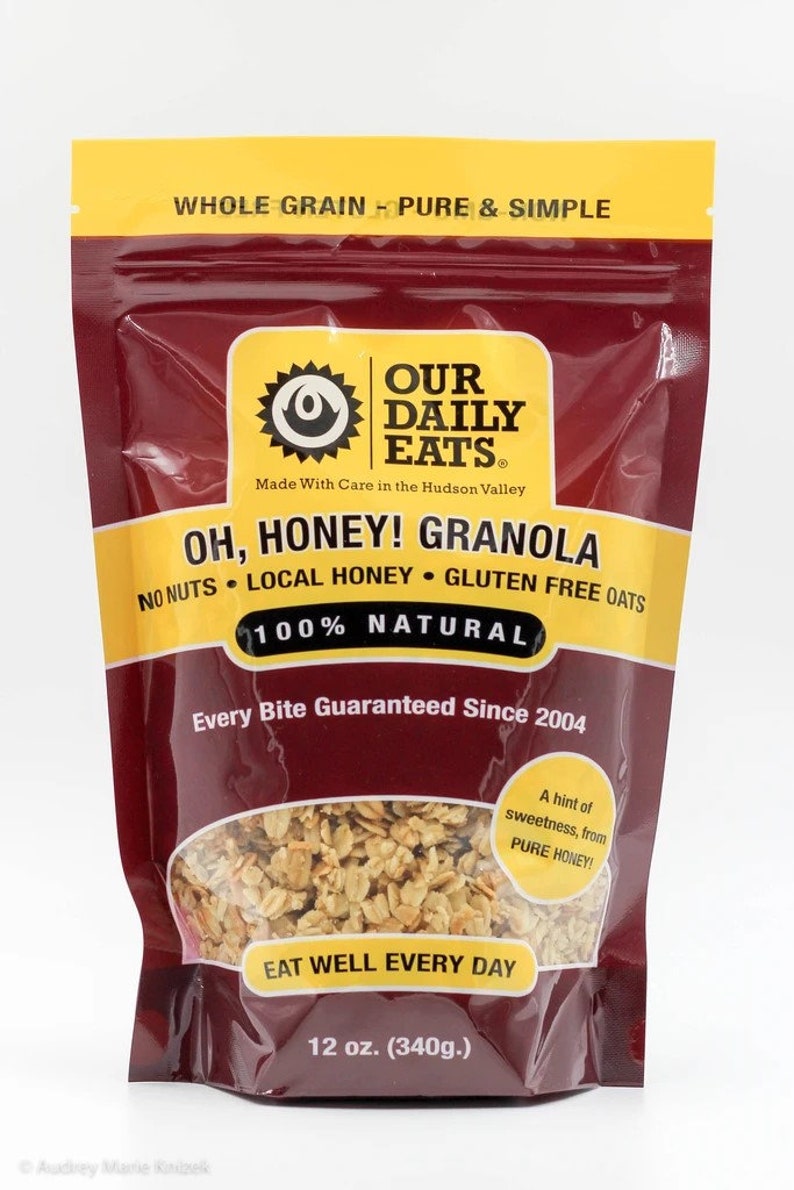 Puede incluir: Una bolsa roja, amarilla y blanca de Our Daily Eats Oh, Honey! Granola. La bolsa presenta un sol con una cara y el texto "Our Daily Eats" en un c&iacute;rculo amarillo. La bolsa tambi&eacute;n presenta el texto "Oh, Honey! Granola" en letras grandes y negritas. La bolsa es de 12 onzas (340 gramos).