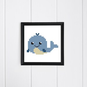 May include: A framed cross-stitch of a blue whale with a light beige belly. The whale has a smiling face and is set against a white background. The frame is black and square, and the artwork is displayed on a white, vertical-paneled wall.