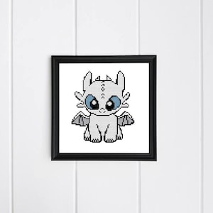 Puede incluir: Un punto de cruz enmarcado de un dragón blanco con grandes ojos azules y pequeñas alas. El dragón está centrado en un marco negro, sobre un fondo blanco.