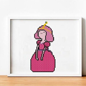 May include: A framed cross-stitch of a cartoon character in a pink dress and crown. The character has pink hair and a smiling face. The artwork is displayed in a white frame, resting on a wooden surface.