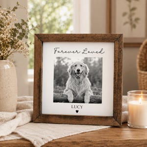 May include: A square wooden picture frame containing a black and white photograph of a golden retriever dog resting its paws on a log. The text Forever Loved is printed above the photo, and the name LUCY with a small heart is printed below.
