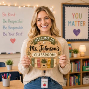 May include: A wooden classroom sign with the text "WELCOME TO Ms. Johnson's CLASSROOM" is held by a smiling person. The sign features decorative elements like leaves, flowers, and books. The background includes a whiteboard with text and a poster that says "YOU MATTER".