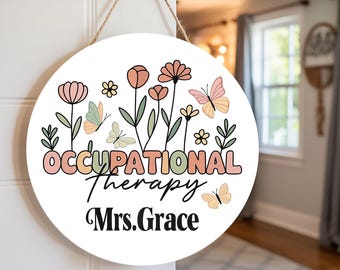 May include: A round white sign with the words "OCCUPATIONAL THERAPY Mrs. Grace" in a decorative font. The sign features floral and butterfly illustrations in pastel colors, with a jute rope for hanging.