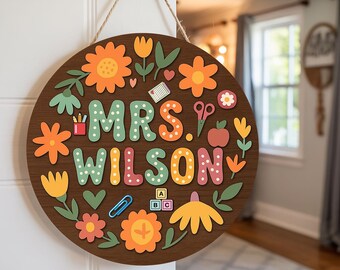 May include: A round wooden sign with the name "MRS. WILSON" in colorful letters, surrounded by floral and school-themed illustrations. The sign is brown with orange, yellow, green, and pink accents, and is hanging by a natural rope.