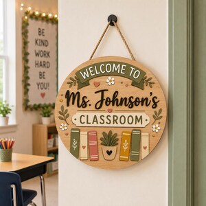 May include: A round wooden classroom sign with the text "WELCOME TO Ms. Johnson's CLASSROOM". The sign has a rope hanger and features decorative elements like books, plants, and floral accents. The sign is hanging on a white wall.