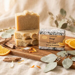 May include: A stack of three beige bars of soap next to a wrapped bar of soap with the text "Soap Citrus Cedarwood Orange Bergamot". The image also includes orange slices, wood pieces, and greenery on a wooden board.