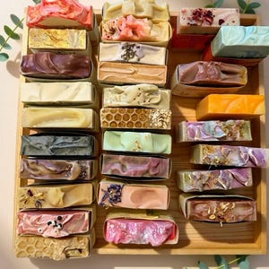 Peut inclure: Un plateau en bois présente un assortiment de savons faits à la main. Les savons rectangulaires affichent diverses couleurs et textures, avec des motifs tourbillonnés, floraux et en nid d'abeilles. Certains sont emballés dans du papier. De la verdure ajoute une touche naturelle.