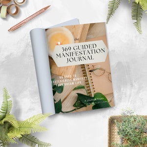 May include: A light blue journal with the title "The 369 Guided Manifestation Journal" and the subtitle "99 Days to Strategically Manifest Your Dream Life". The journal is open to reveal a page with a gold spiral binding and a quote from Kimberley Lowe.