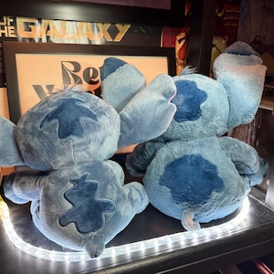 May include: Two blue plush toys featuring large ears and dark blue patches on the back and head. The figures are seated side by side, showing their backs. Each plush has a soft, textured surface and a small tail. The toys are positioned on a shelf.