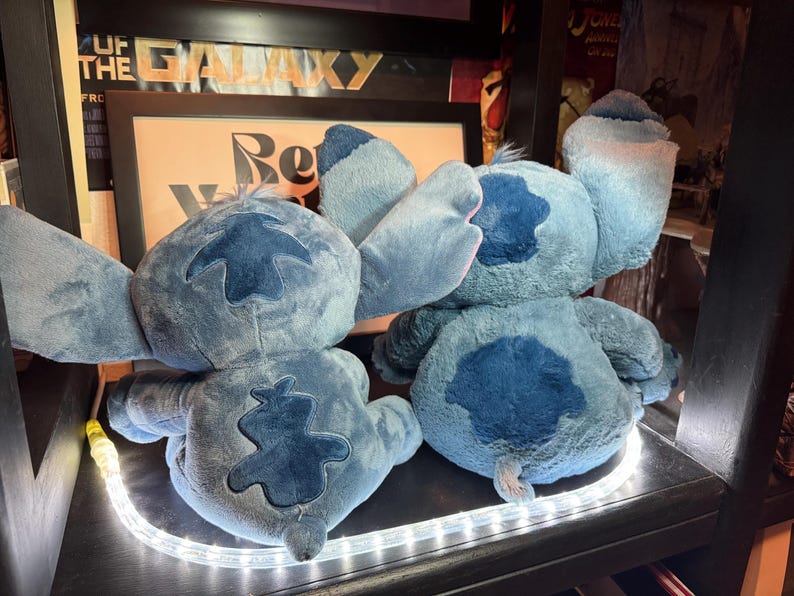 May include: Two blue plush toys featuring large ears and dark blue patches on the back and head. The figures are seated side by side, showing their backs. Each plush has a soft, textured surface and a small tail. The toys are positioned on a shelf.
