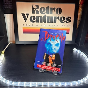 May include: A paperback copy of Bram Stoker's Dracula, a novel and film adaptation, with a black and white photo of the main character on the cover. The book is on a shelf with a white string of lights.