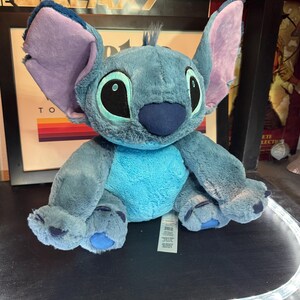 May include: A plush toy of the character Stitch, featuring soft blue fur, a lighter blue chest patch, large pointed ears with lavender inner lining, and oversized black eyes with light blue irises. The stuffed animal has a dark blue nose and paws.