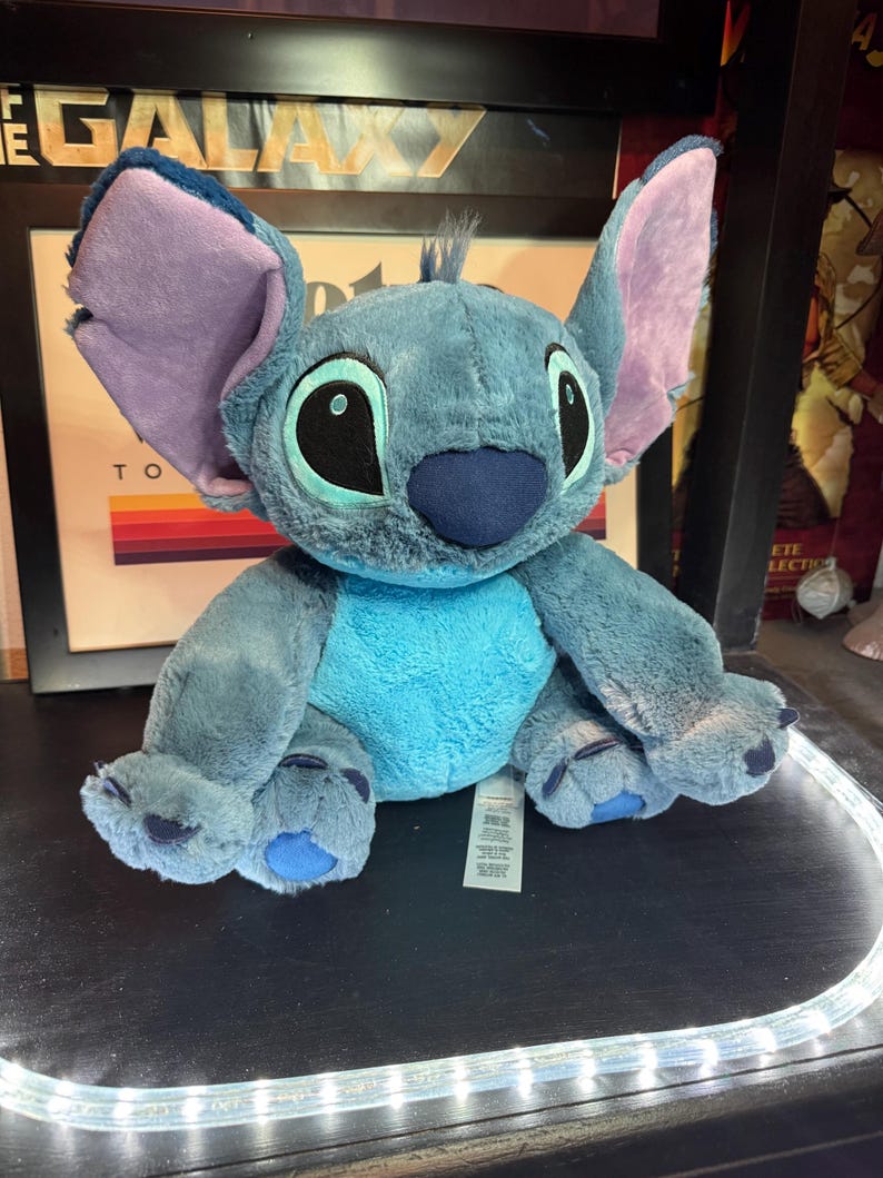 May include: A plush toy of the character Stitch, featuring soft blue fur, a lighter blue chest patch, large pointed ears with lavender inner lining, and oversized black eyes with light blue irises. The stuffed animal has a dark blue nose and paws.