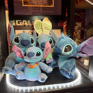 May include: A collection of four blue plush toys depicting the character Stitch. Three figures are seated, with one wearing a floral lei and another wearing yellow and blue polka-dot bunny ears. The fourth figure is positioned behind the others.