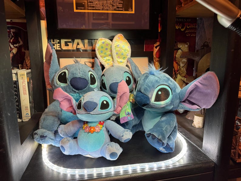 May include: A collection of four blue plush toys depicting the character Stitch. Three figures are seated, with one wearing a floral lei and another wearing yellow and blue polka-dot bunny ears. The fourth figure is positioned behind the others.